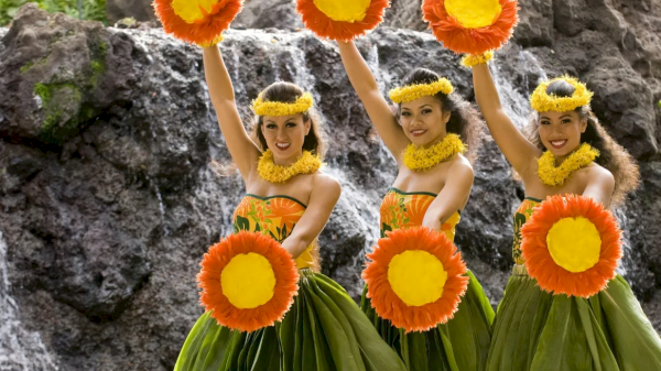 Three dancers in vibrant costumes hold large flower props, showcasing a festive and colorful performance near a rocky backdrop.