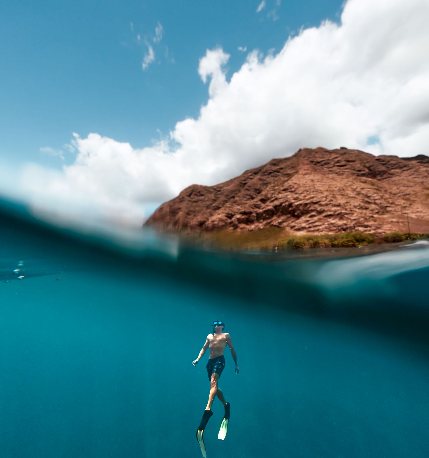 A person is snorkeling in clear blue water, with a rocky shore and clouds in the background, showcasing a vibrant underwater scene.