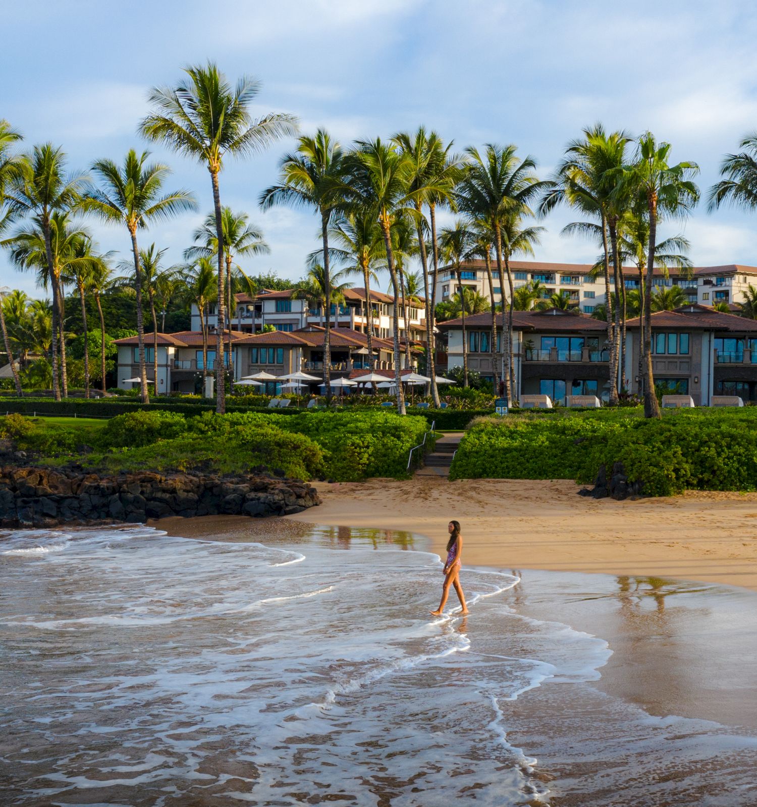 A serene beach scene with a person walking along the shore, palm trees, and a luxurious resort in the background.