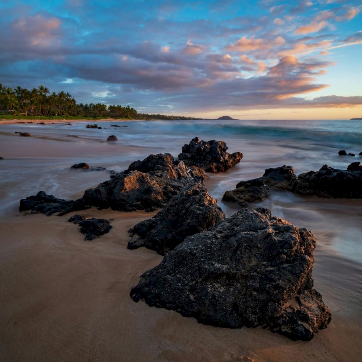 The image shows a coastal scene with rocks on the beach, gentle waves, and a colorful sky at sunset. It captures serene beauty.
