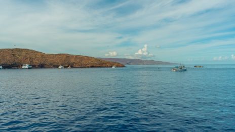 The image shows a serene coastal view with calm blue waters, distant land, and boats, under a clear sky with scattered clouds.