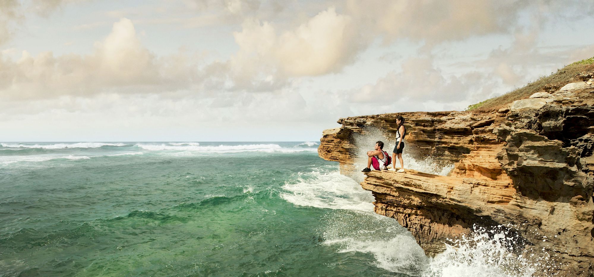 The image shows a rocky cliff overhanging a turbulent ocean, with two people sitting near the edge and waves crashing below.