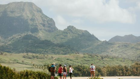 A group of people stands near a scenic landscape with mountains and greenery, enjoying nature's beauty on a sunny day.
