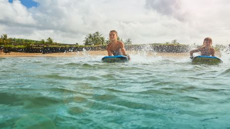 Two kids ride bodyboards in the ocean, kicking up water as they enjoy a sunny day at the beach. It's a scene of summer fun.