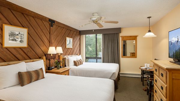 The image shows a cozy hotel room with two beds, wooden accents, a TV, and a balcony overlooking nature. It&rsquo;s inviting and warm.