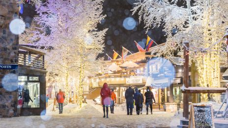 A snowy night scene features cheerful people walking amid glowing trees and festive lights, creating a magical winter atmosphere.