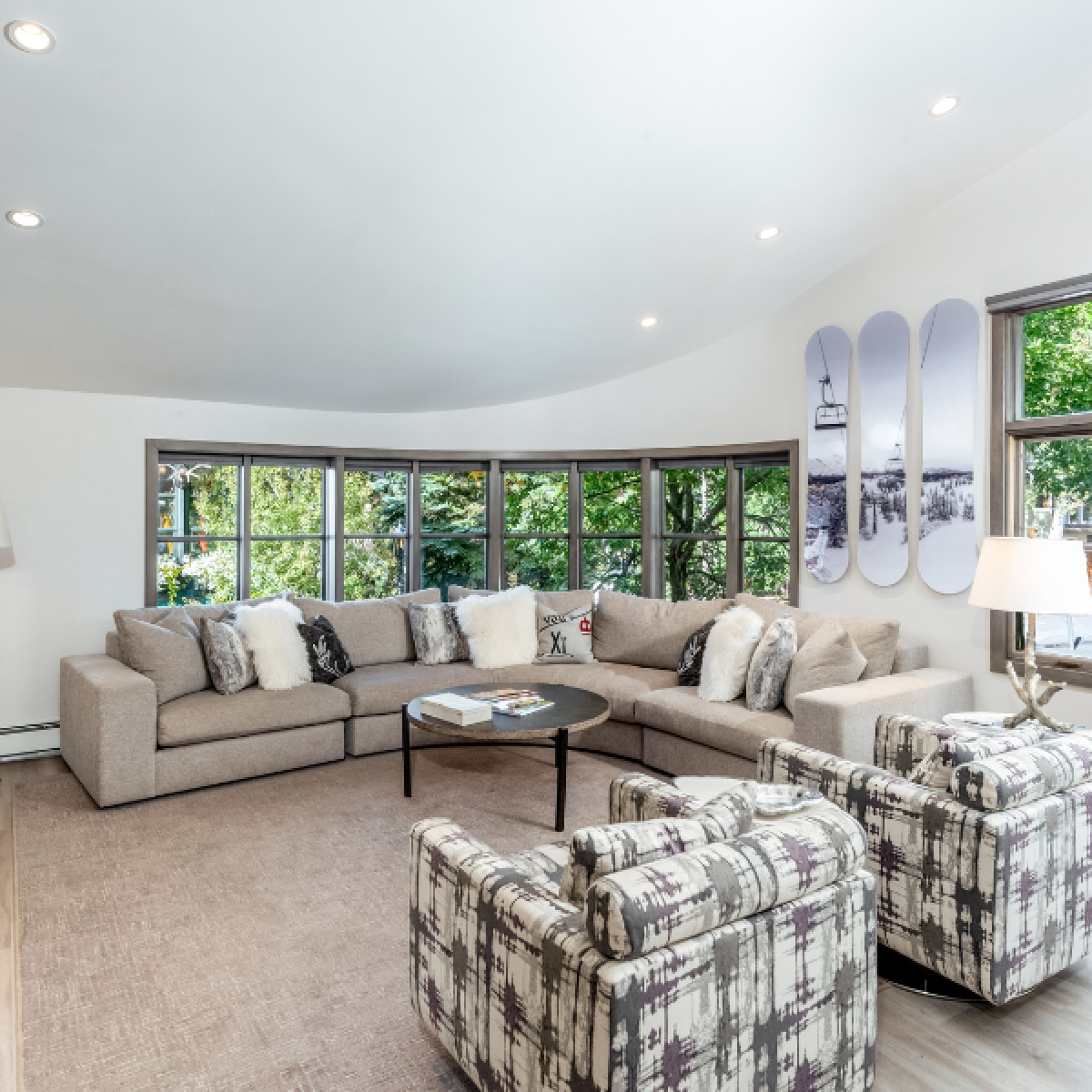 The image showcases a cozy living room with a sectional couch, patterned chairs, a coffee table, and large windows inviting natural light.