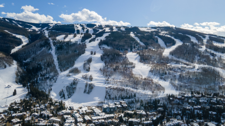 The image shows a snowy mountain landscape with ski slopes, framed by a clear blue sky and a village at the base.