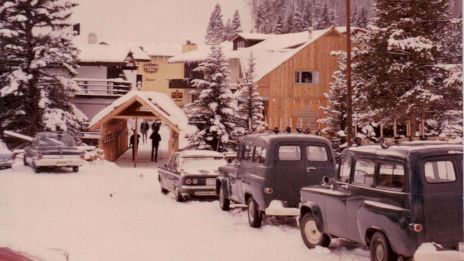 The image shows a snowy mountain resort with parked cars, a ski slope in the background, and people walking towards the lodge.