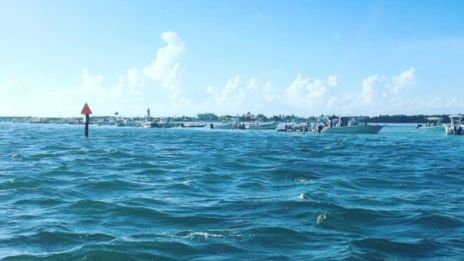 The image shows a calm water scene with boats in the distance, under a bright blue sky with scattered clouds.