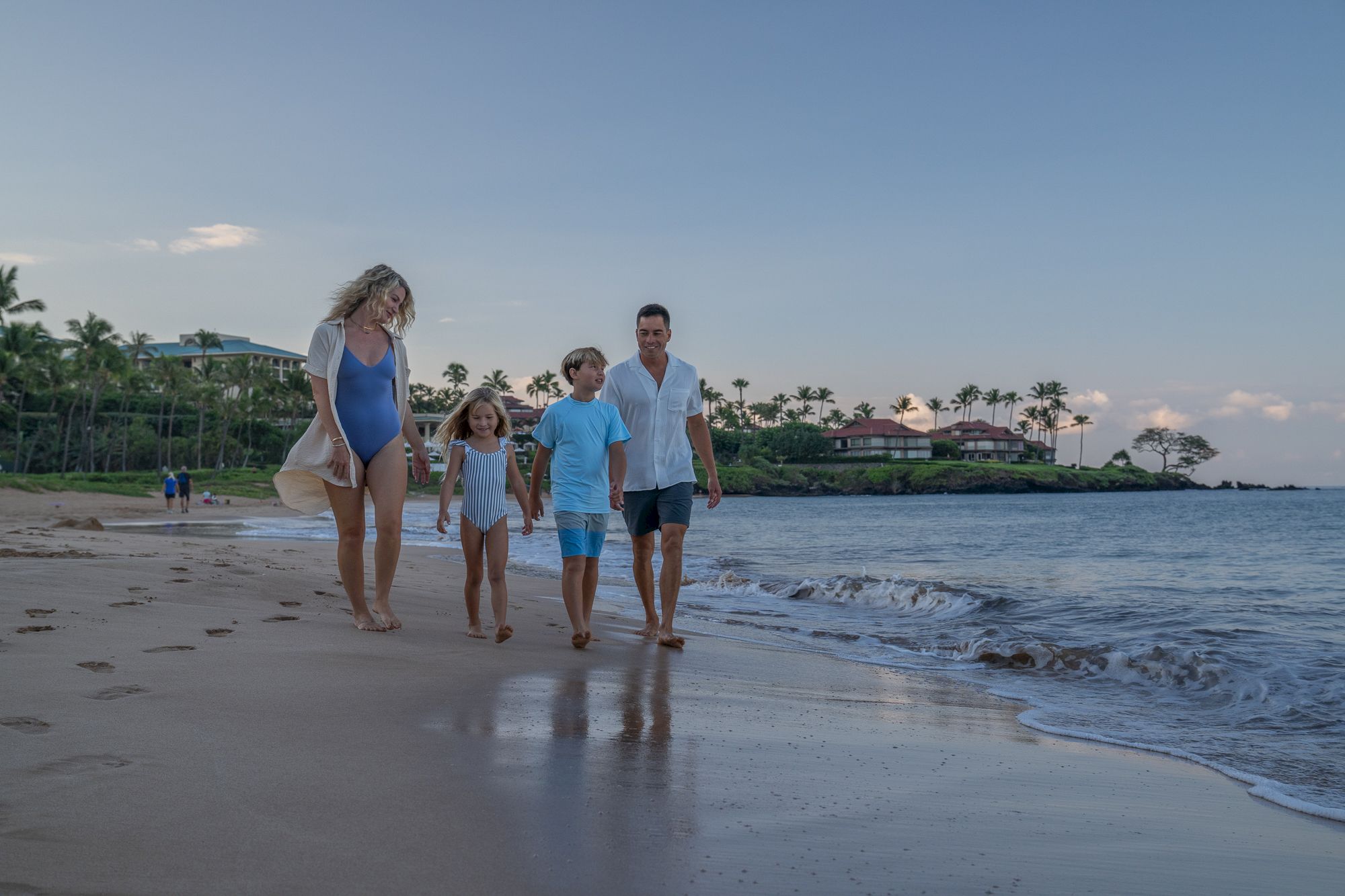 A family walks along a sunny beach at the edge of the water, holding hands and smiling, with palm trees and resort buildings in the background.