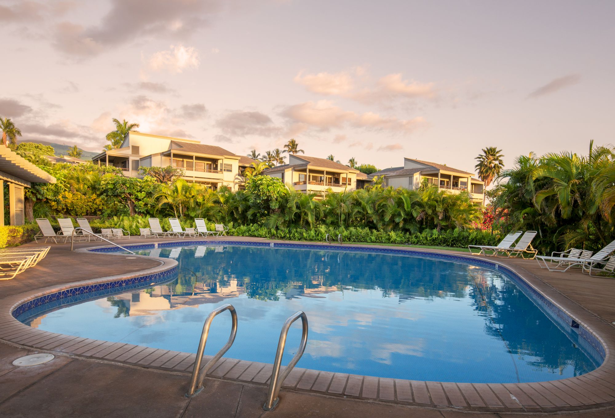 The image shows a serene pool area surrounded by greenery and buildings, reflecting a peaceful atmosphere in the evening light.