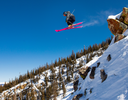 A skier launches off a snowy cliff on a bright blue day, mid-air above a rocky, pine-covered slope.