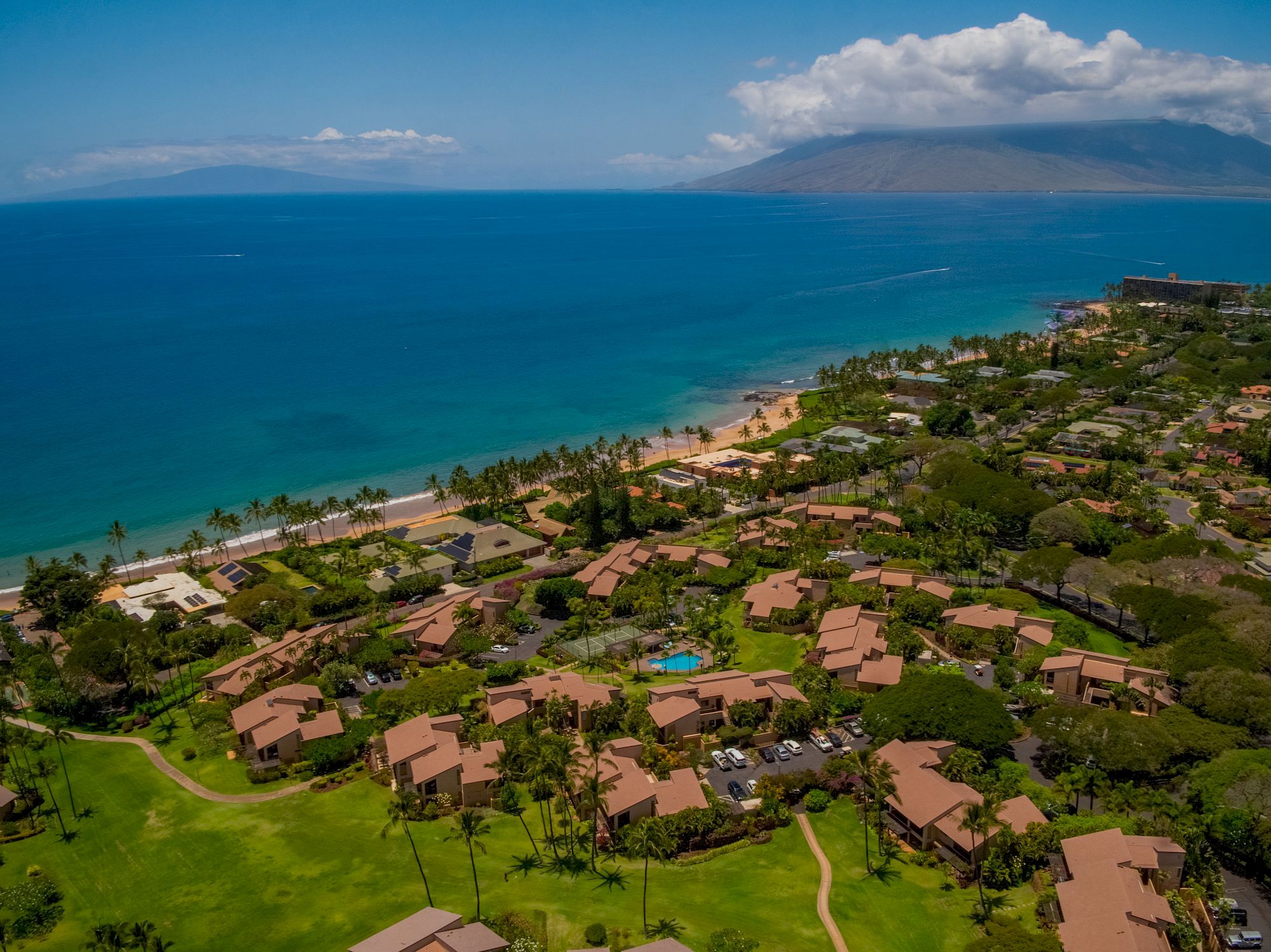 The image shows a coastal landscape with lush greenery, beachfront properties, and a clear blue ocean under a sunny sky.