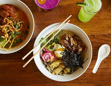 Two ramen bowls on a wooden table with toppings like seaweed, bamboo shoots, fish cake, and pickled greens; chopsticks rest on the bowls.