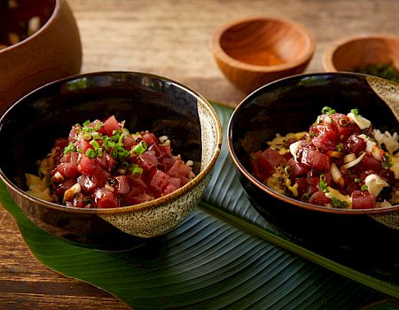 Two bowls of poke/tuna over rice, diced tuna, cilantro, onions, and seasonings on a wooden table with green leaves.