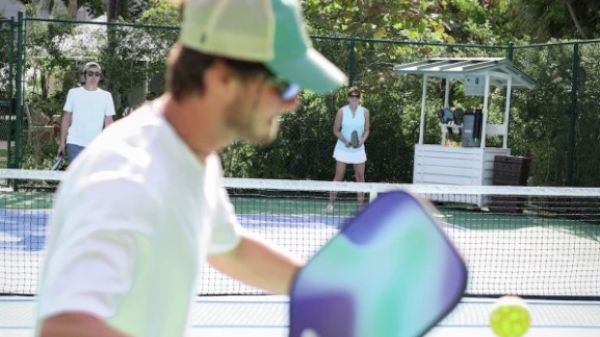 The image shows a man playing paddle tennis, with a woman in the background and another person observing nearby.