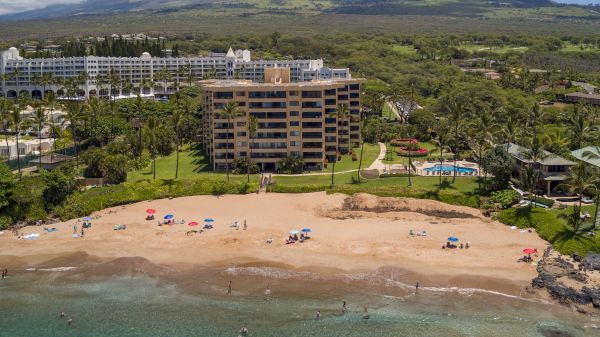 Aerial view of a sandy beach with umbrellas, people, a nearby hotel, lush greenery, and mountains in the background.