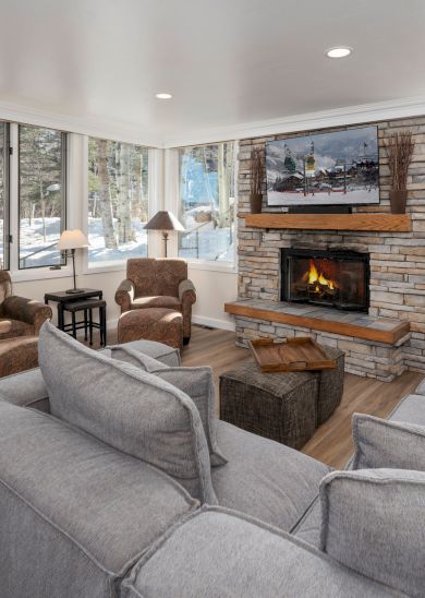A cozy living room with a fireplace, gray sofas, armchairs, and large windows showing a snowy landscape, decorated with a wall art piece.