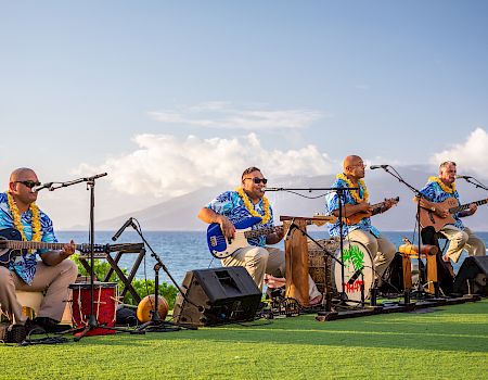 A band plays acoustic instruments outdoors by the ocean, wearing Hawaiian shirts and leis, with audio equipment set up.