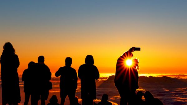 Silhouetted people watch a vibrant sunset, with one person taking a photo, capturing the sunburst over a cloud-filled landscape.