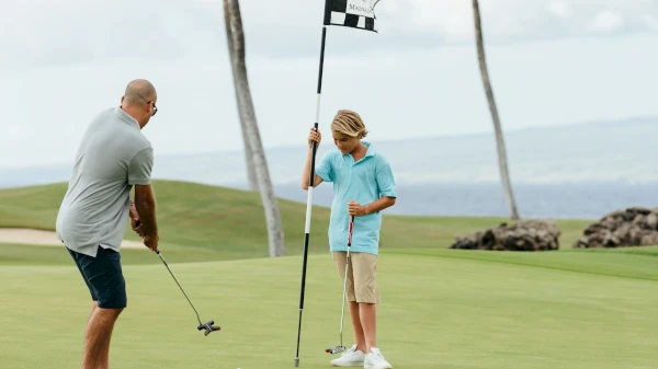 A person is putting on a golf course while another holds the flagstick. The setting is outdoors with a scenic view in the background.