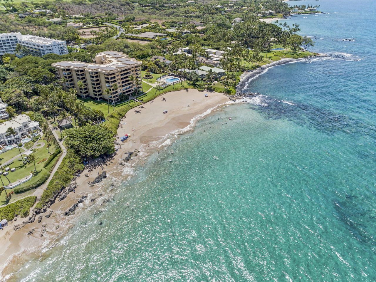 Aerial view of a coastal area with clear blue water, sandy beach, and green vegetation alongside buildings and a developed area.