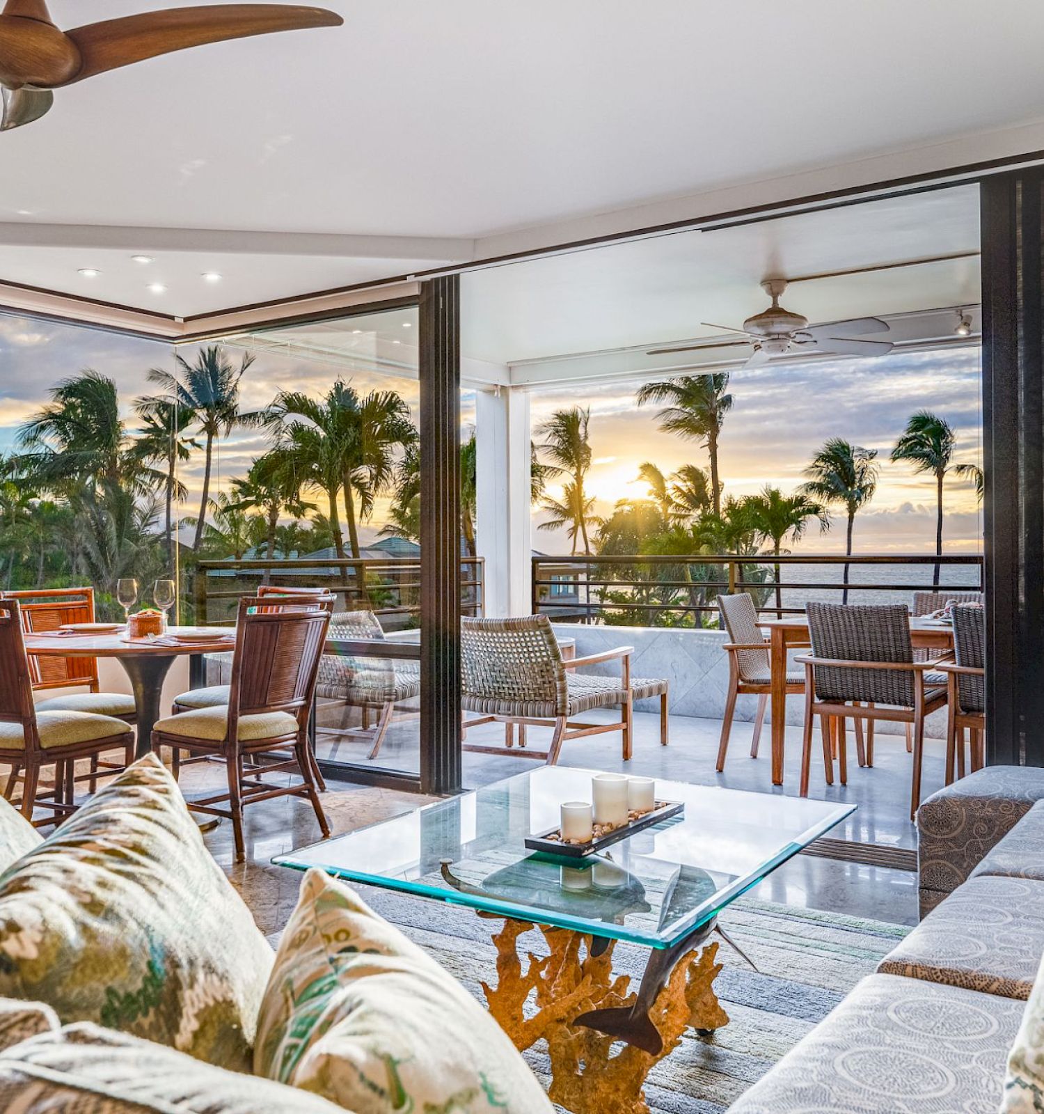 Spacious living room with modern decor, large glass doors open to a patio with dining area, offering a stunning view of a tropical sunset.