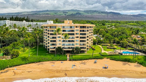 A beachfront building with lush greenery, a pool, and people on the beach; mountains and cloudy skies in the background.