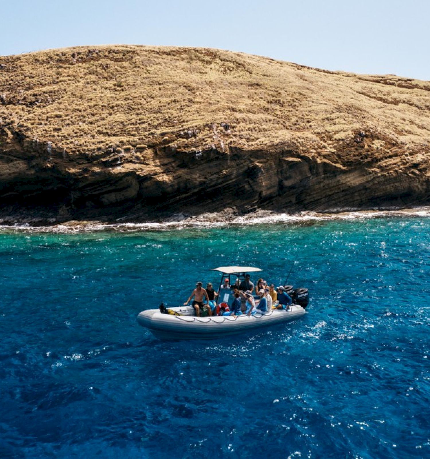 A small boat with people on it navigates blue waters near a rocky coastline under a clear sky.