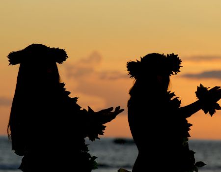 Two silhouetted dancers wearing leafy headpieces and arm garlands perform against a sunset backdrop over the ocean.