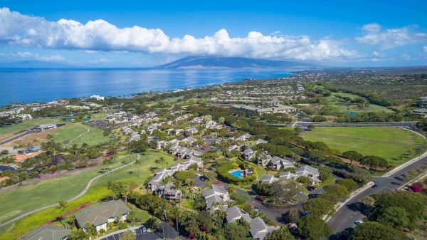 The image shows a coastal landscape with residential areas, greenery, and ocean views under a clear blue sky with clouds.