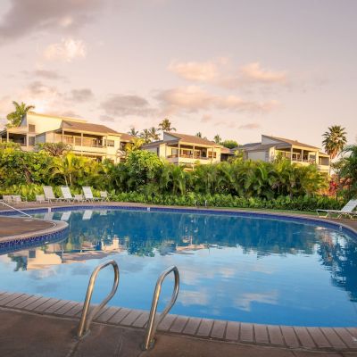 The image shows a tranquil pool surrounded by greenery and lounge chairs, set against a serene sky at dusk.