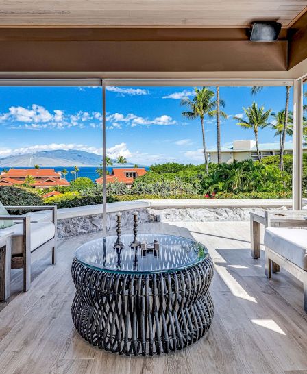 The image features a stylish outdoor seating area with plush chairs, a unique table, and a stunning view of the ocean and palms.