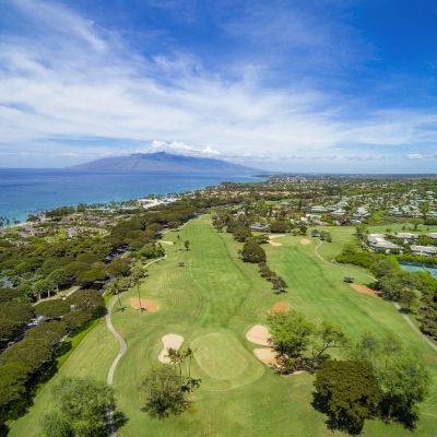 The image shows a scenic golf course with lush greenery, ocean views, and distant mountains under a bright sky. Beautiful setting.