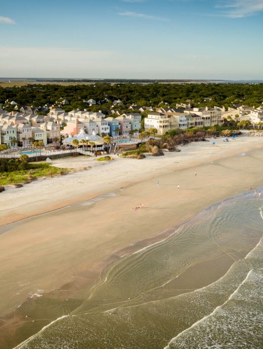 A beachfront scene with a long sandy beach, waves, and a row of houses beside dense greenery under a blue sky.