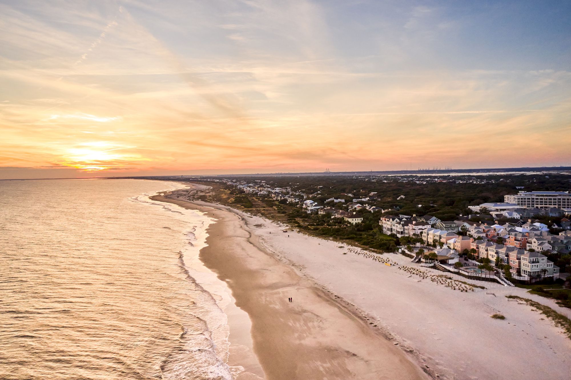 A coastal view at sunset showing a sandy beach, ocean waves, and nearby buildings under a colorful sky.