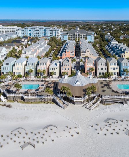 Aerial view of a beachfront resort with colorful houses, two swimming pools, and white sandy beach with numerous umbrella stands visible.