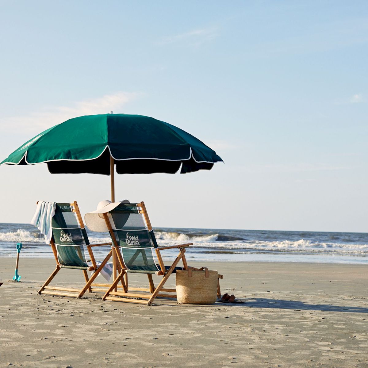 Two beach chairs under a green umbrella face the ocean on a sandy beach, with a small basket and calm waves in the background.