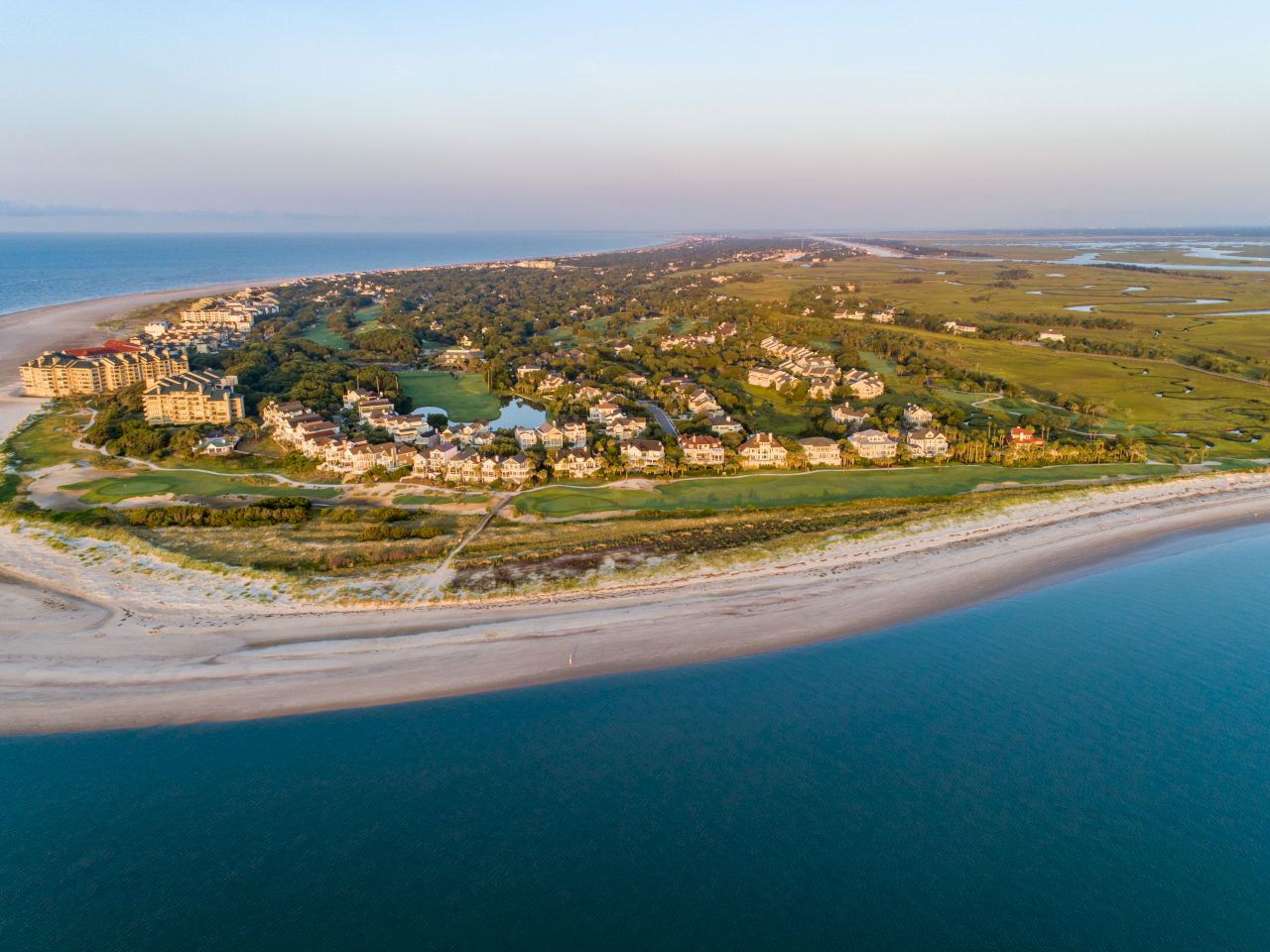 A coastal aerial view features a strip of sandy beach, residential houses, and lush greenery meeting a tranquil blue sea.
