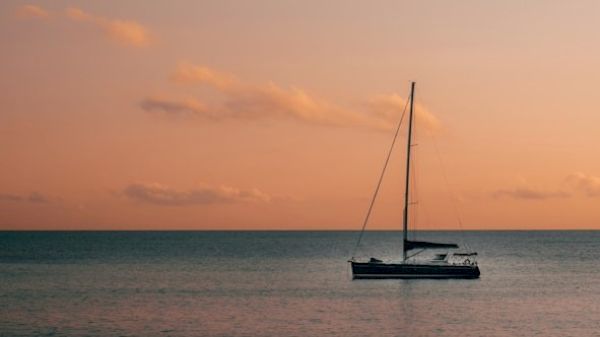 A solitary sailboat floats on calm waters under a pastel-colored sunset sky with scattered clouds.