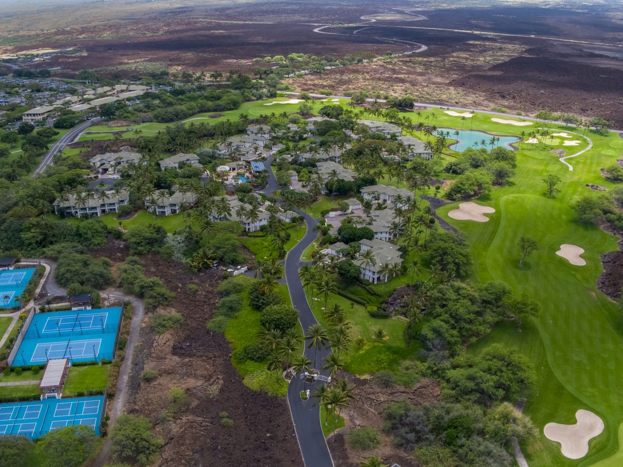 Aerial view of a lush area with a golf course, residential buildings, tennis courts, and winding roads surrounded by greenery and lava rocks.