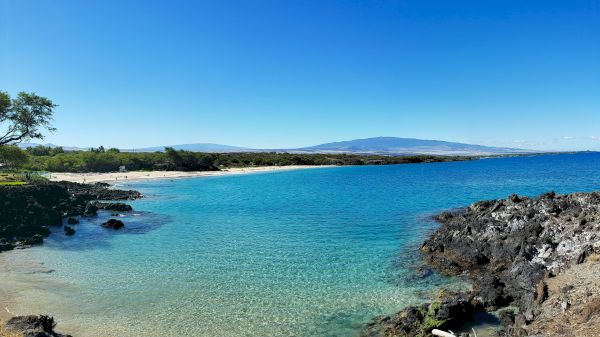 A beautiful beach scene with clear turquoise water, rocky coastline, and a mountainous backdrop under a bright blue sky.