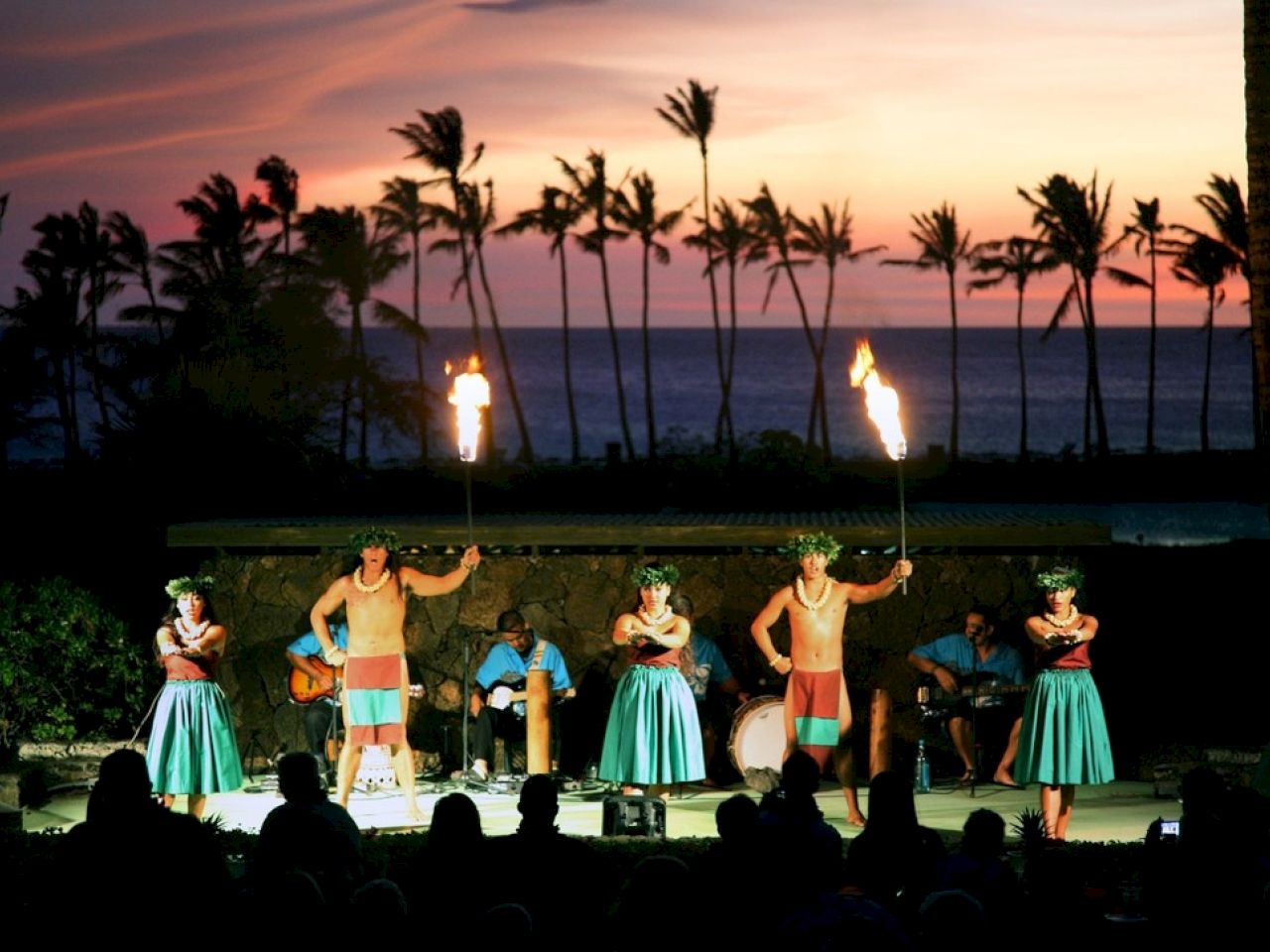 A Hawaiian luau featuring dancers with torches, musicians, and a backdrop of ocean and palm trees during sunset.