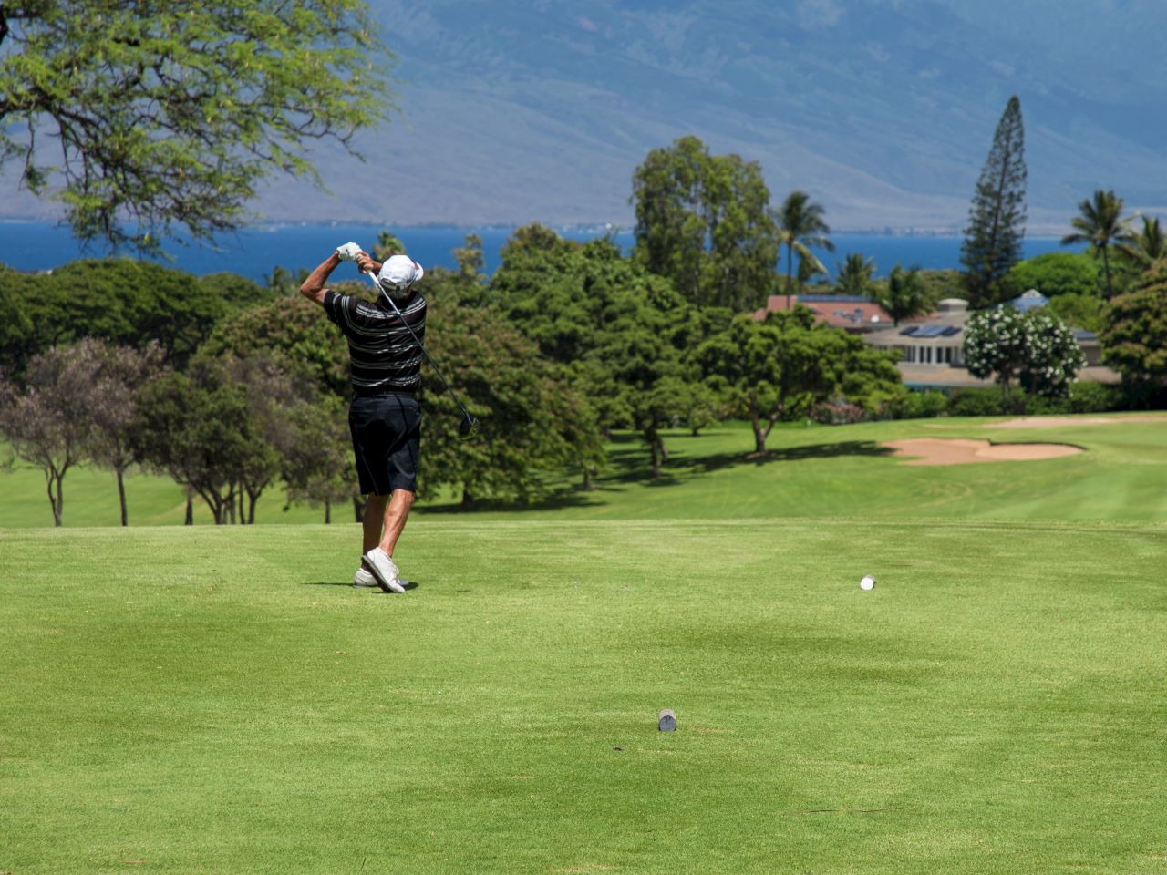 A person is playing golf on a green course with mountains and trees in the background under a partly cloudy sky.