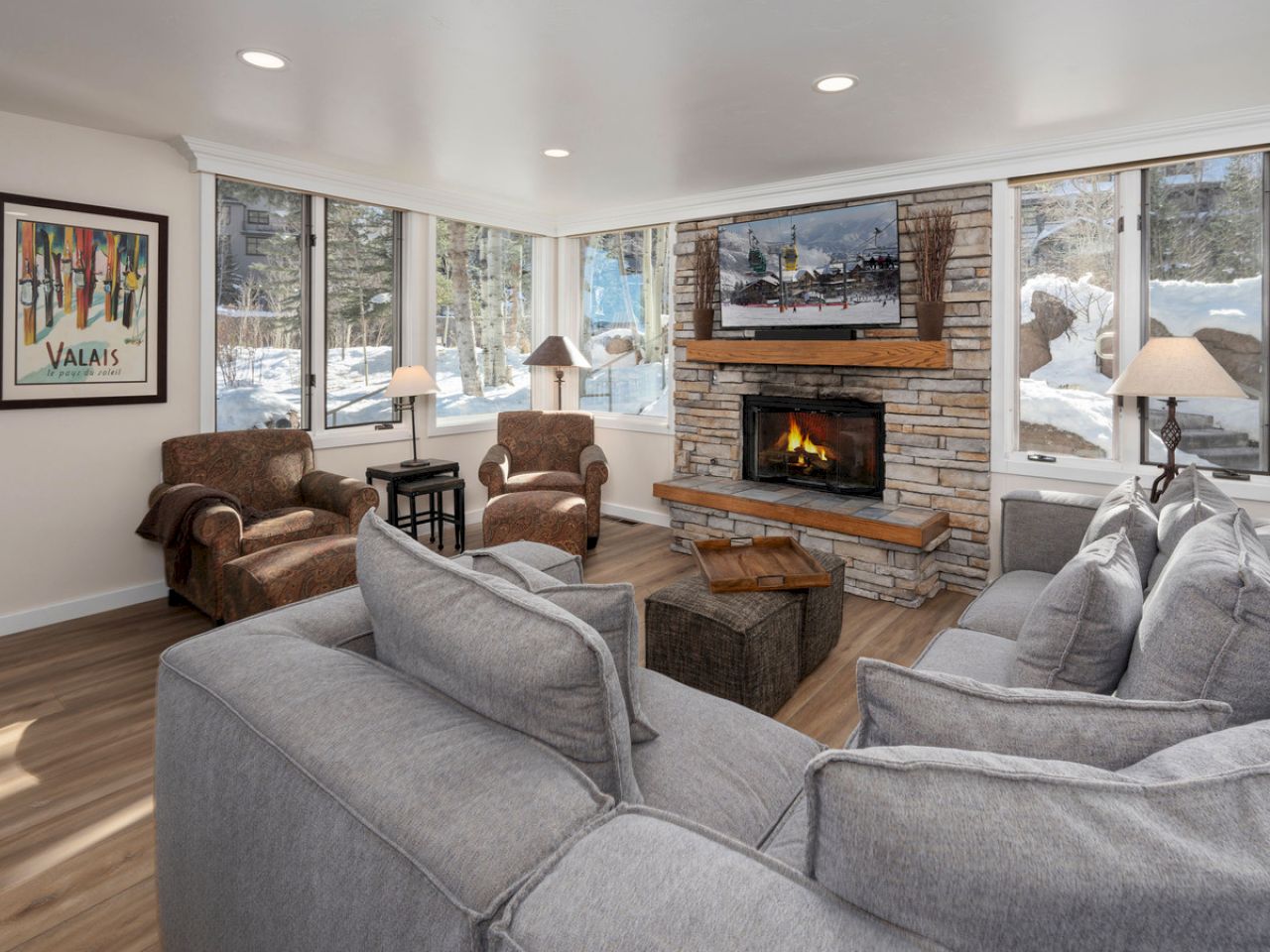 A cozy living room with gray sofas, armchairs, a stone fireplace, and large windows with snowy views. A poster titled "Valais" is on the wall.
