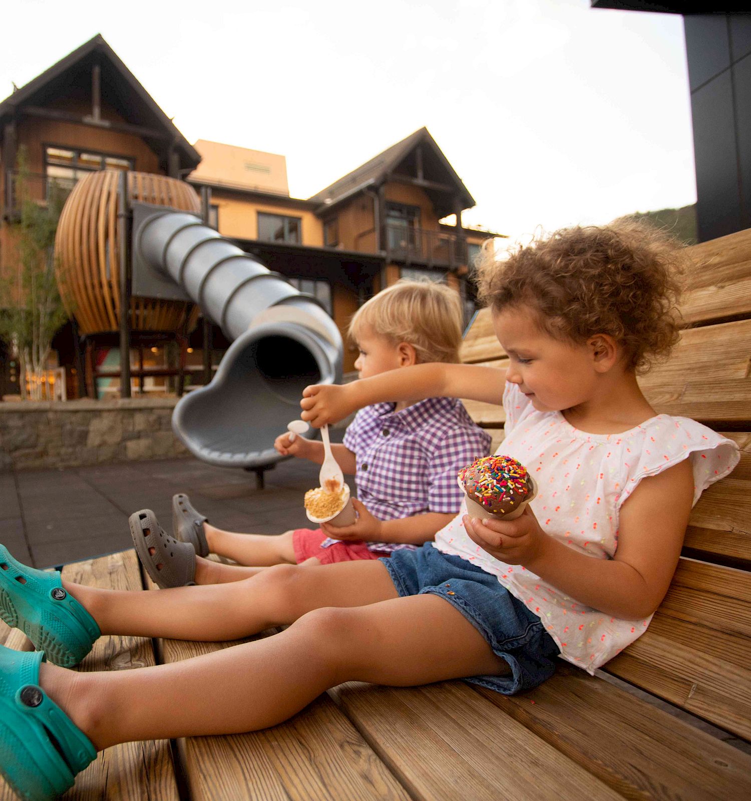Two children sitting on a wooden bench enjoying ice cream cones, with a large slide in the background.