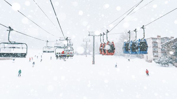 A snowy ski resort scene with people on ski lifts and skiers on the slopes; surrounding buildings are visible through the falling snow.