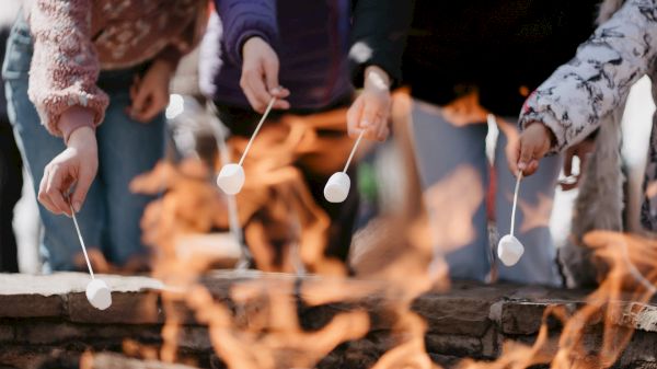 People roasting marshmallows over an open fire, holding them on sticks, creating a warm and cozy outdoor gathering atmosphere.
