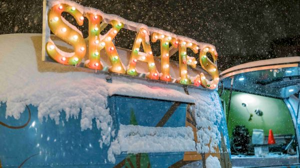 A snow-covered trailer with a brightly lit "SKATES" sign on top, set against a snowy backdrop, likely at an ice skating venue.